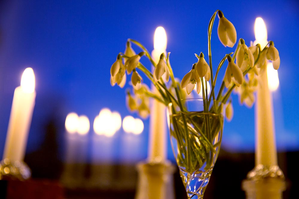 Snowdrops in a glass and lit candles on a windowsill, reflections of the candles can be seen in the glass and through the window the evening sky is a deep blue